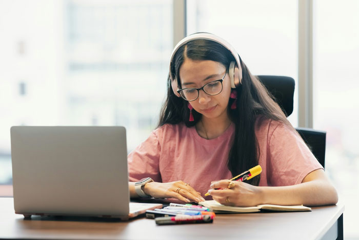 young woman studying on a desk with a laptop nearby, she's highlighting text on a book and is wearing headphones